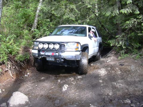 1991 Ford Bronco - A white SUV navigates a challenging, muddy off-road trail surrounded by dense forest foliage.