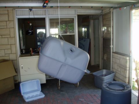 1990 Ford Bronco - A gray fuel tank is shown hanging from a rope inside a room, with various containers and boxes nearby.