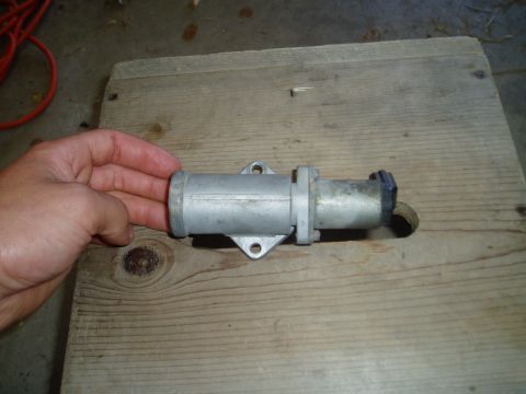 1990 Ford Bronco - A hand holds a cast-aluminum idle air control valve against a weathered plank of wood on a garage floor, showing its two-bolt mounting flan…