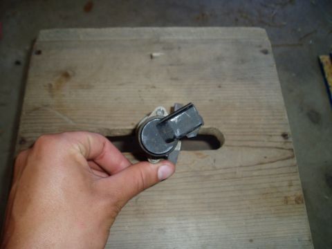 1990 Ford Bronco - A hand holds a small used idle air control valve over a plywood bench top, showing its round body, gray plastic two-pin connector housing a…