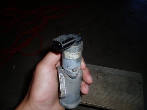 1990 Ford Bronco - A hand holds a used Ford idle air control (IAC) valve with its black two-pin connector on top and a paper label reading "FOTE-9A" with a da…