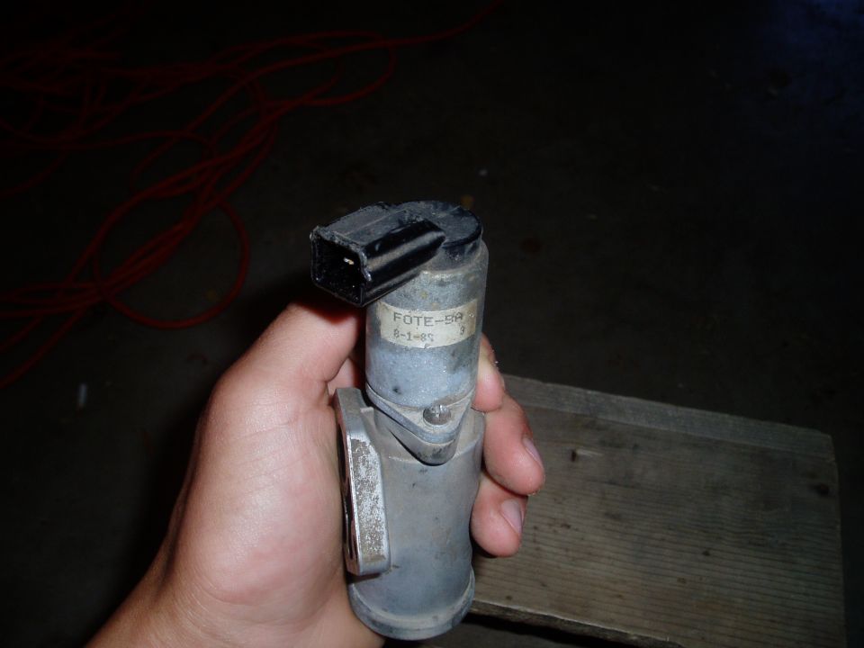 Hand holding a used Ford idle air control valve labeled FOTE-9A over a garage floor