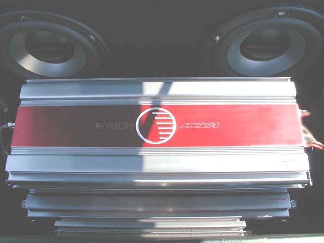 Car audio amplifier with red stripe and "ORION HCCA" logo, stacked on another amplifier, with two subwoofers behind