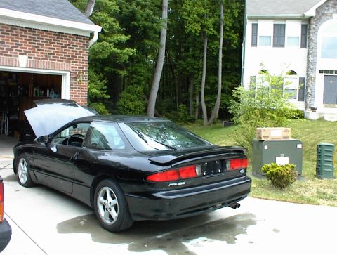1994 Ford Probe - A black 1994 Ford Probe is parked in a driveway with its hood open, next to a garage and a house.