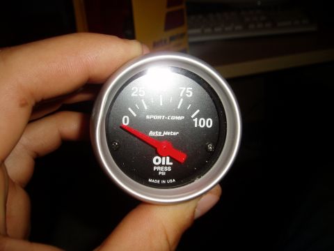 1990 Ford Bronco - A hand holds a new AutoMeter Sport-Comp oil pressure gauge, black face with a 0-100 PSI scale, red pointer resting at zero and a brushed-si…