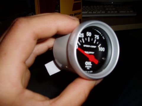 1990 Ford Bronco - A hand holds a new Auto Meter Sport-Comp oil pressure gauge with a black face, 0-100 PSI scale and red pointer in a silver bezel. The Auto…