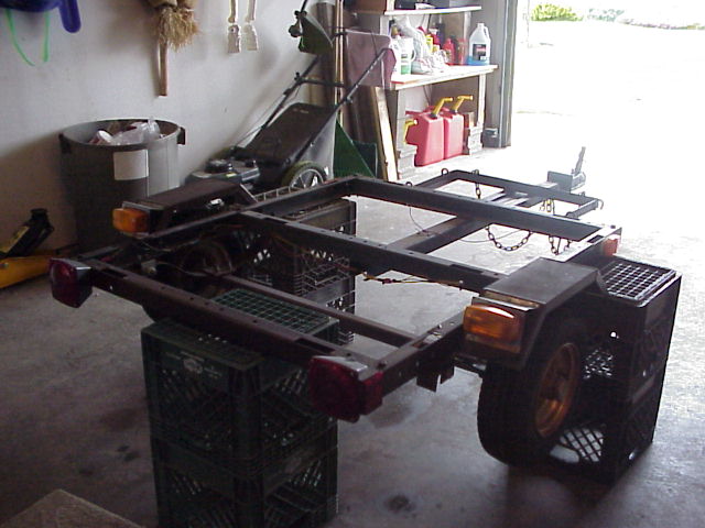 Bare trailer frame with tail lights propped on milk crates in a garage, one wheel visible