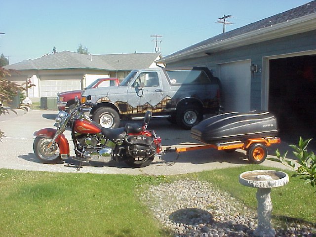 Red Harley cruiser towing a small trailer in a driveway, black lifted Bronco behind