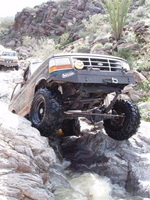 1994 Ford Bronco - A dark-colored 1994 Ford Bronco tackles a challenging rocky stream crossing, with a second Bronco visible on the trail behind it.