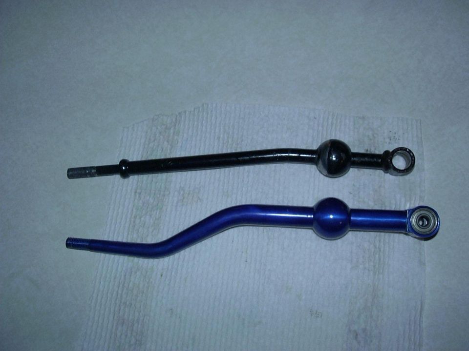 Two shifter levers, one black and one blue, lie on a white textured surface. The blue lever has a bearing visible at its end