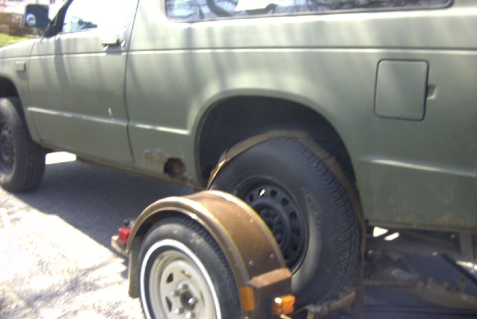 Olive-green compact pickup with a spare wheel and brake drum resting against it