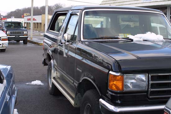 Black full-size Ford Bronco with chrome side steps parked in a shop lot, snow on cowl