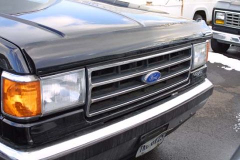1989 Ford Bronco - Close-up of the front end of a black late-1980s Ford truck or Bronco, showing the chrome-bar grille with blue Ford oval, rectangular sealed…