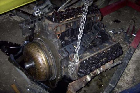 1989 Ford Bronco - A greasy V8 engine block sits on a shop floor with the intake manifold and cylinder heads' valve covers removed, exposing the pushrods, roc…