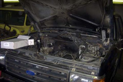 1989 Ford Bronco - Open engine bay of a dark blue 1980s Ford truck in a garage with the radiator, fan shroud and much of the front-end plumbing removed, expos…