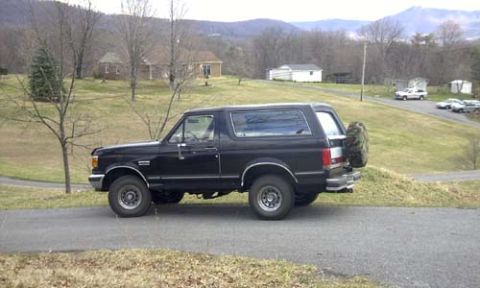 1989 Ford Bronco - Side profile of a black late-1980s Ford Bronco with its removable hardtop in place and a covered spare tire mounted on the tailgate, parked…
