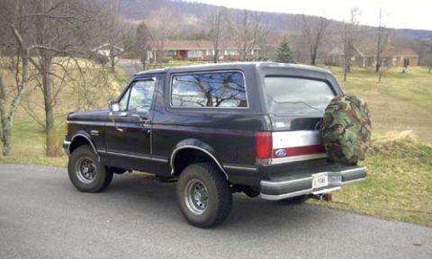 1989 Ford Bronco - Driver-side three-quarter rear view of a black full-size Ford Bronco with a silver bodyside stripe and a camouflage-print cover on the tail…