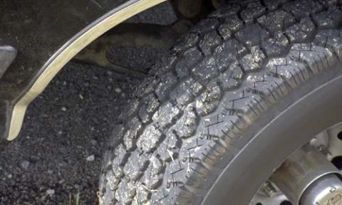 1989 Ford Bronco - Tight close-up of an all-terrain tire's tread and shoulder blocks on a truck parked on gravel, with bits of stone and shiny debris lodged b…