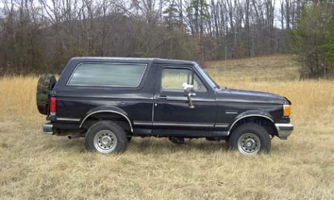 1989 Ford Bronco - Side profile of a black late-1980s Ford Bronco parked in tall dry grass, showing the removable hardtop with large side glass, a camouflage-…