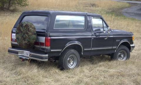 1989 Ford Bronco - Rear three-quarter view of a black late-1980s Ford Bronco in a dry grass field, with a camouflage-covered spare tire on the tailgate, chrom…