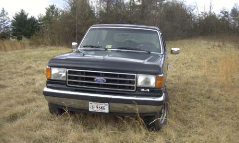 1989 Ford Bronco - Head-on view of a black late-1980s Ford Bronco parked in tall dry grass, showing the blue Ford oval in the chrome egg-crate grille, amber c…