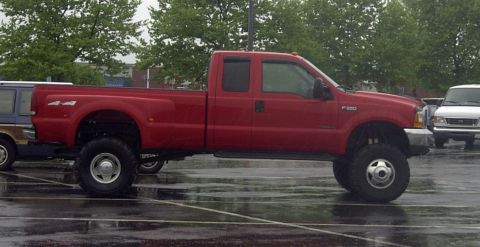 1989 Ford Bronco - Side profile of a lifted red Ford F-350 Super Duty extended-cab dually pickup on oversized mud tires, parked on a wet parking lot under gre…