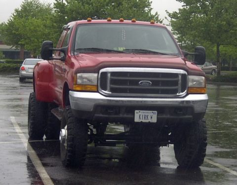 1989 Ford Bronco - Front view of a lifted red Ford Super Duty dually pickup on tall mud tires in a rainy parking lot, showing the chrome grille with blue Ford…