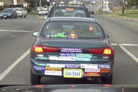 1989 Ford Bronco - Rear view of a dark green Ford Escort sedan stopped in traffic, its trunk lid and bumper plastered with dozens of slogan bumper stickers in…