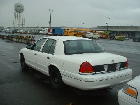 1999 Ford Crown Victoria - A white sedan is parked on a wet lot with a water tower in the background.