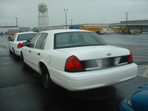 1999 Ford Crown Victoria - Two white Ford Crown Victoria police cars are parked in a lot under a cloudy sky. A water tower and school buses are visible in the backgro…