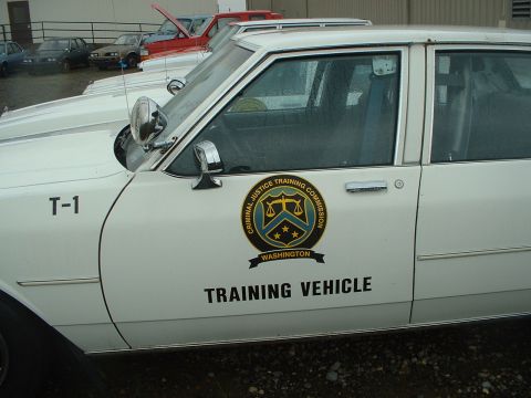 1999 Ford Crown Victoria - A white sedan, marked as a training vehicle for the Criminal Justice Training Commission, is parked in a lot with other vehicles.