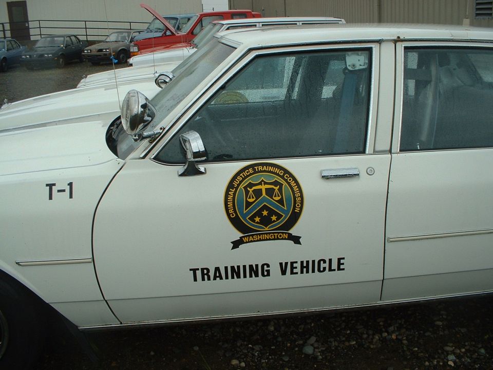 White sedan side view, "T-1" on door, "Criminal Justice Training Commission Washington" emblem, "TRAINING VEHICLE" text