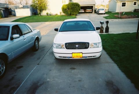 2000 Ford Crown Victoria - A white 2000 Ford Crown Victoria is parked in a driveway, with another car to its left and a garage in the background.