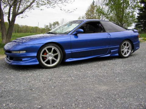1994 Ford Probe - A blue coupe with aftermarket wheels is parked on a gravel surface, with trees and foliage in the background.