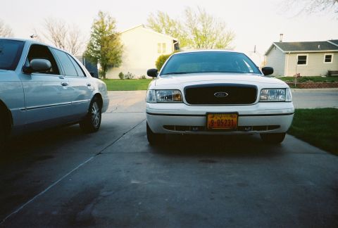 2000 Ford Crown Victoria - Two sedans are parked in a driveway, one white Ford Crown Victoria and a silver car.