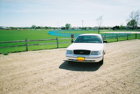 2000 Ford Crown Victoria - A white 2000 Ford Crown Victoria is seen from a 3/4 front view, parked on a gravel lot next to a fenced-in sports field.