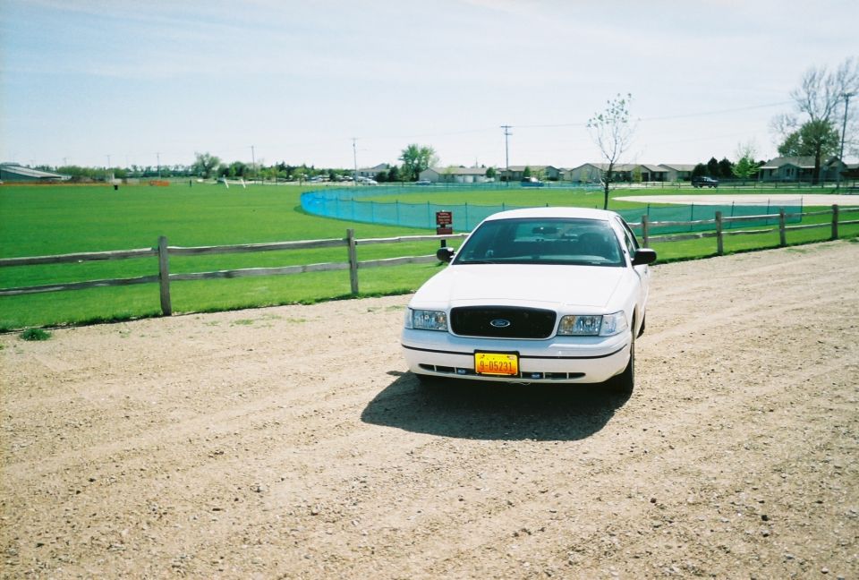 White 2000 Ford Crown Victoria parked on a dirt road near a sports field