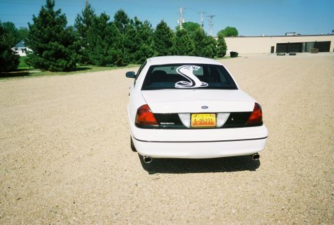 2000 Ford Crown Victoria - A white sedan is parked in a gravel lot with trees and a building in the background.