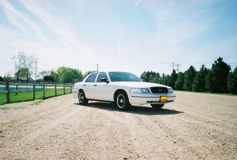 2000 Ford Crown Victoria - A white 2000 Ford Crown Victoria is parked on a gravel road with a wooden fence and trees in the background.