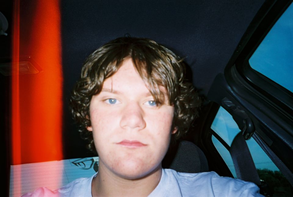 Person with curly hair and blue eyes inside a car, looking at the camera. Red light streaks on the left