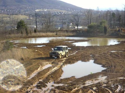 1989 Ford Bronco - A lone green Jeep Wrangler with a hardtop sits in the middle of a wide red-clay mud flat crossed with tire ruts and standing water. A town…