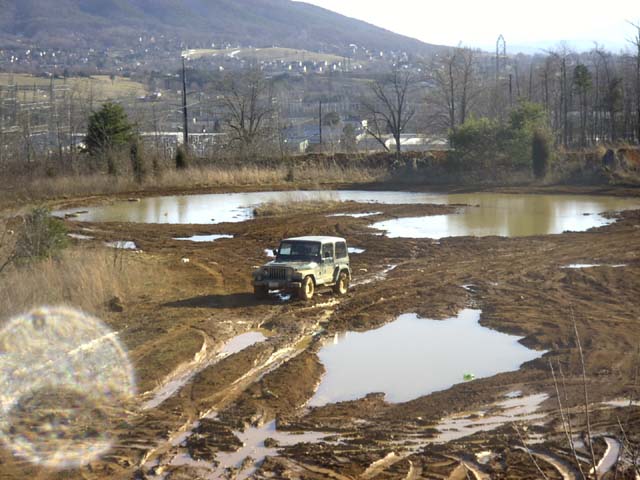 Green hardtop Jeep Wrangler parked on a rutted clay mud flat with water pools