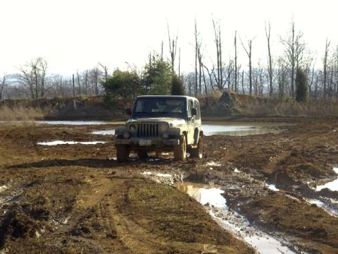 1989 Ford Bronco - A Jeep Wrangler with a light-colored hardtop sits nose-on in a churned mud pit, its round headlights and seven-slot grille facing the camer…