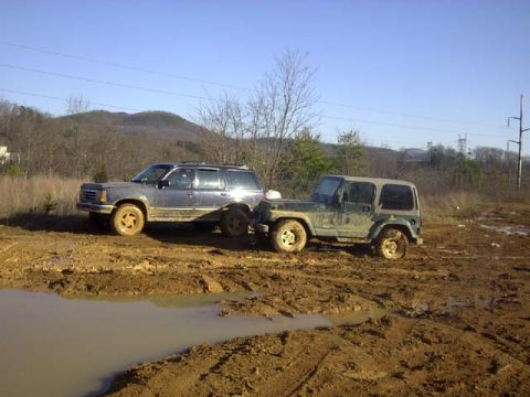 1989 Ford Bronco - A mud-caked dark blue Ford Explorer four-door SUV nose-to-tail with a green Jeep Wrangler in a churned red-clay mud pit, both riding on whi…