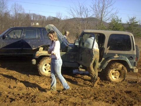 1989 Ford Bronco - A green Jeep Wrangler with its hood up and driver's door open sits stuck in deep red-clay mud while people stand around it, one leaning int…