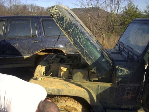 1989 Ford Bronco - A mud-caked dark green Jeep Wrangler with its hood propped open in a field, exposing the engine bay, coolant reservoir and radiator hose; t…