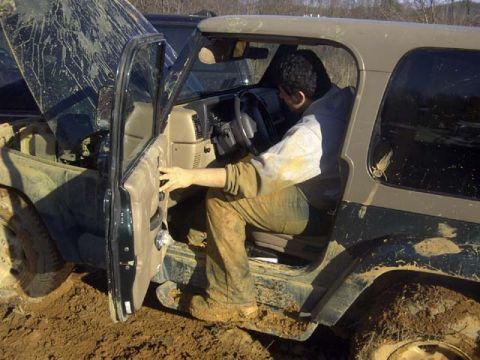 1989 Ford Bronco - A person in mud-splattered clothes leans into the driver's seat of a dark green Jeep Wrangler with a tan hardtop, the door swung open to sh…