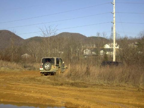 1989 Ford Bronco - A Jeep Wrangler with a tailgate-mounted spare tire sits on a rutted orange-clay mud trail cut through dry winter brush, with standing water…
