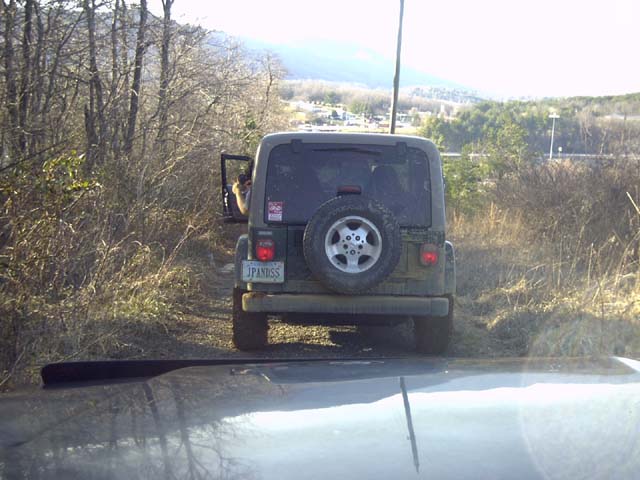 Rear view of a muddy green Jeep Wrangler with spare tire on a brushy trail