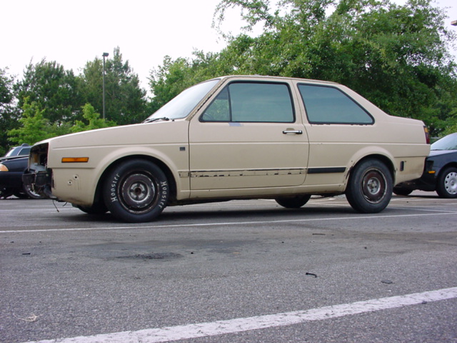 Tan two-door sedan with racing tires and missing front bumper. Appears to be a 1984 Volkswagen Golf/Jetta